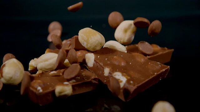 SLOW MOTION, MACRO, DOF: Nuts and chocolate chips scatter over a few squares of milky chocolate. Blanched hazelnuts and bittersweet cocoa drops fall over a pile of milk chocolate with hazelnuts.
