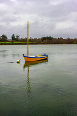 a small yellow sailboat at anchor. A sailboat is at anchor, the current is quite strong. The weather is cloudy, the coast is visible.