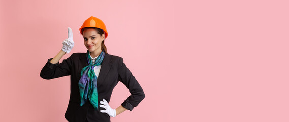 Happy young girl, female flight attendant in orange color protective helmet posing isolated on pink studio background.