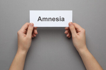 Woman holding white card with word Amnesia on grey background, top view