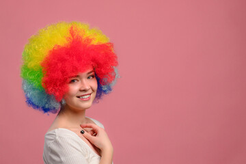 Smiling young attractive female in colorful wig looks at camera on pink background. Close-up.