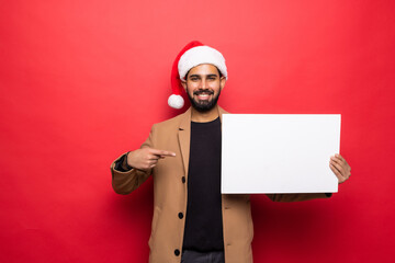 Young indian man in santa hat holding white blank board standing on red background