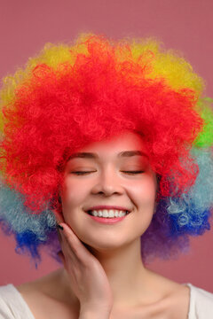 Smiling young attractive female in colorful wig with closed eyes on pink background. Close-up.