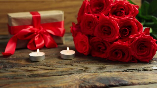 celebrating Valentine's Day holiday concept: wrapped git box with red ribbon bow, ruby roses bouquet and burning candles on wooden table. romantic greetings love woman, birthday or dating presents