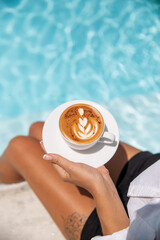 Image of Young caucasian woman drinking cappuccino with latte art closed to blue swimming pool
