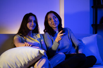 Fototapeta na wymiar two young women watching tv and chatting laughing, women watching tv with a remote at home at night laughing