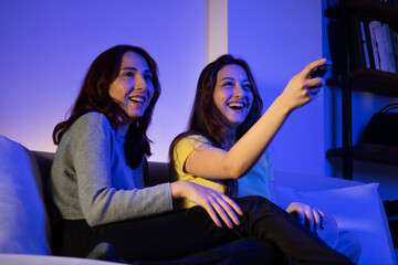 two female friends watching tv laughing at home, women watching tv with a remote at home at night laughing