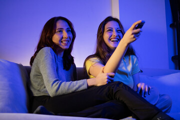 two female friends watching tv laughing at home, women watching tv with a remote at home at night laughing