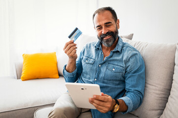 Man with beard using credit card to make online payment on tablet PC. Man holding debit card and...