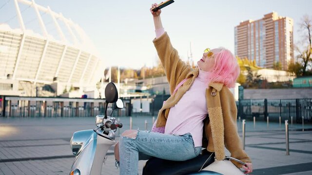 Young positive woman with pink hair making selfie on smartphone, posing on scooter at parking area, tracking shot