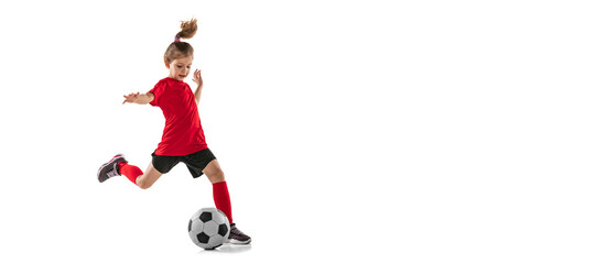 Full-length portrait of sportive child, girl in red uniform playing, training football isolated...