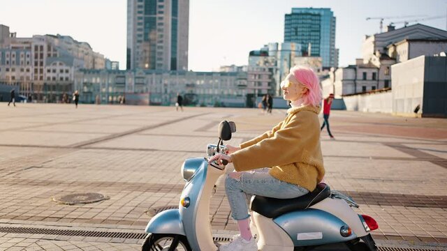 Young stylish woman with pink hair riding on scooter along city square, enjoying good weather , tracking shot