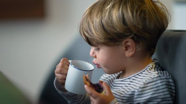 Toddler eating bread with jelly and drinking from mug in breakfast