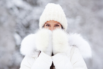 Closeup portrait of attractive woman in white knitted hat, and fur mittens on snow cold winter nature background. Beautiful brown eyes, model covering with hands her mouth outdoor