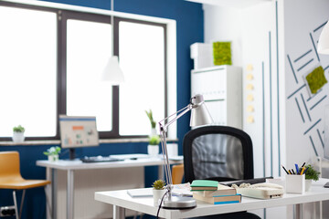 Lamp on a white desk in an empty modern business office. Startup workspace furniture with blue and white wall. Table in commercial workplace with big window. Nobody in indoors work location.