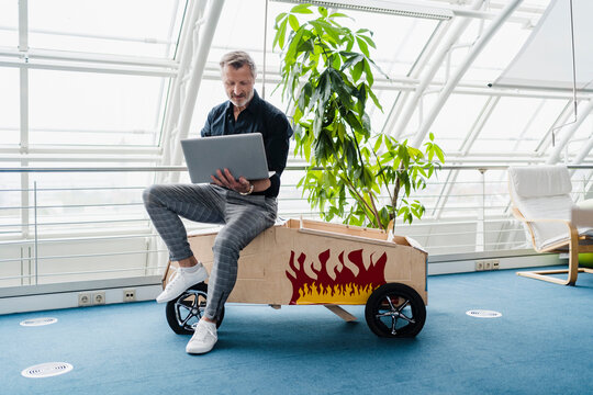 Businessman using laptop while sitting on toy car in creative office
