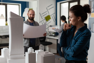 Architect in modern office thinking about design work standing next to tall building foam model with team colleague engineer holding blueprints. Team of architects doing creative thinking.