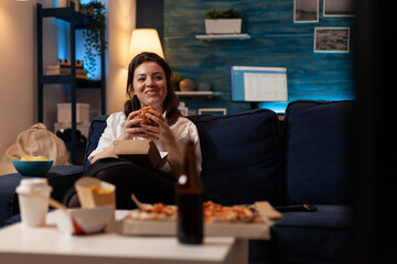 Happy woman eating tasty takeaway hamburger relaxing on couch watching comedy television series in living room. Smiling person having takeout burger and fast food delivery menu.