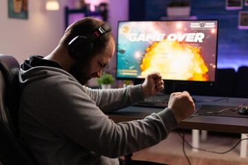 Disappointed man losing video games on computer. Adult clenching fists and playing games with headphones and joystick, feeling sad about lost game. Player using modern equipment.