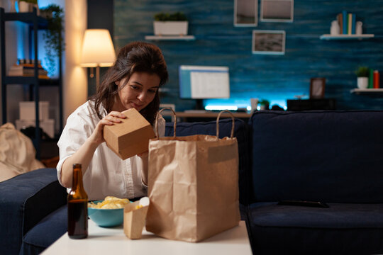 Woman On Sofa Unpacking Burger Box From Paper Takeaway Bag Sitting On Couch In Front Of Table With Beer Bottle, Fries And Bowl Of Chips. Person In Living Room Unpacking Fast-food Home Delivered.