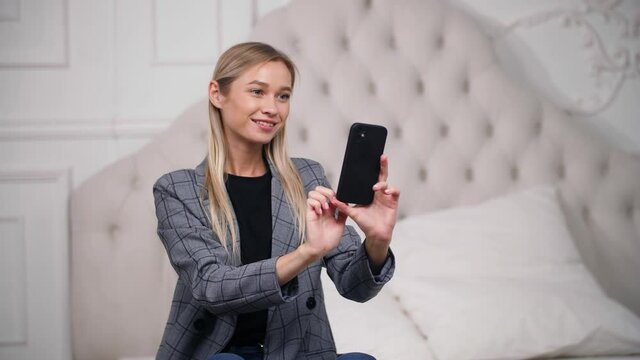 Young adult joyful female poses on bed in gray coat to take cute pictures, photos with phone for social media likes. Trendy woman touches smartphone screen to record, film video of herself in bedroom.