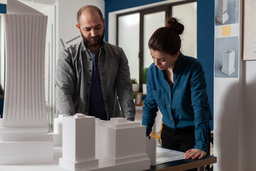 Architectural engineers doing teamwork reading construction plans standing next to table with foam model maquette. Team of two architects colleagues working together looking down at blueprins.