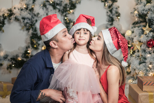 Christmas studio family shoot in red Christmas hats. New Year photo of happy mixed family with cute daughter. Mother and father kissing their daughter