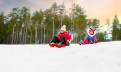 childhood, sledging and season concept - happy little girls sliding on sleds down snow hill in...