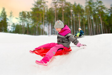 childhood, sledging and season concept - happy little girl sliding down on sled outdoors in winter over snowy park or forest background - obrazy, fototapety, plakaty