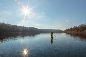 Fototapeta na wymiar Late autumn on a clear day a man is rafting on a supboard on the river. backlight, sun in the frame. SUP. Stand up paddle boarding
