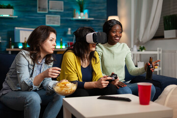 Concentrated woman with virtual reality headset holding controller playing videogames while multi-ethnic friends helping her during online competition enjoying spending time together. Hanging out