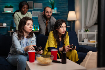 Concentrated women sitting on sofa playing videogames using joystick during online competition enjoying spending time together in living room. Group of multi-ethnic friends having fun at home