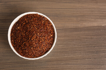 Obraz na płótnie Canvas Dry rooibos leaves in bowl on wooden table, top view. Space for text