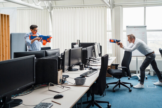 Male colleagues playing with toy gun while standing by desk