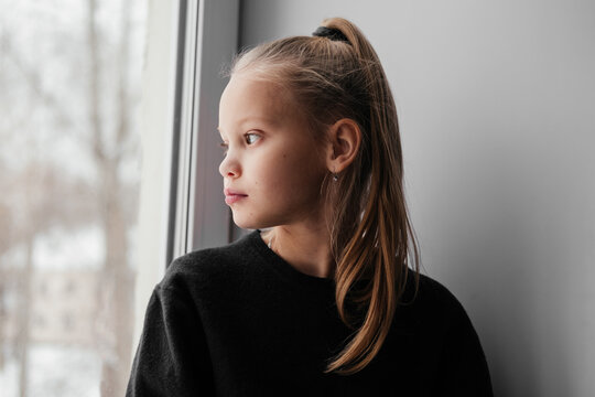 Bored lonely little girl in black sweater staring at window. 10 years old child home alone sitting on window sill and daydreming. Kid looking outside on snowy street