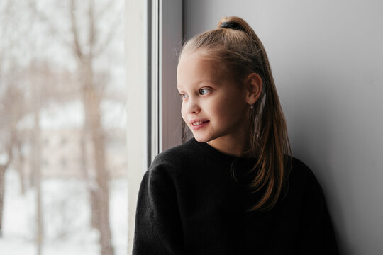 Smiling little girl sitting and watching out of window. 10 years old kid home alone during lockdown, quarantine. Child in sweater waiting for parents staring at window