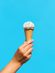 food, dessert and eating concept - close up of hand holding vanilla ice cream in waffle cone with sprinkles over blue background