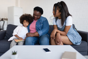 african american girl looking at picture book while sitting on couch with granny and mom