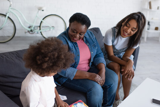 cheerful african american women looking at curly girl sitting on couch with picture book