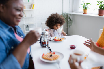 african american child drinking tea during breakfast with blurred granny and mom
