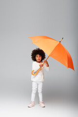 full length view of african american girl standing with orange umbrella on grey