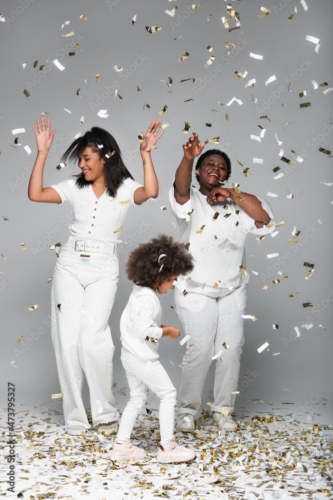 Wall mural happy african american women and child in white clothes under shiny confetti on grey