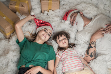 Christmas photo of happy Family. New Year Portrait of dad, mom and daughter wearing Christmas red hats, smiling and laying in white artificial snow. 