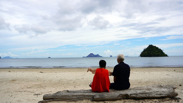 Asian senior couple with mask relax sitting on beach log white sand landscape and beautiful sea