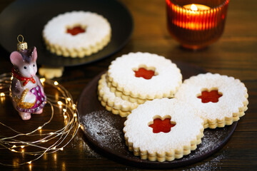 Linzer cookies served on dark plate, Christmas glass decoration, twinkle lights and burning candle. Dark wooden table, golden backdrop, warm light, high resolution