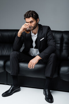 young bearded man in formal wear sitting on black sofa and drinking whiskey on grey