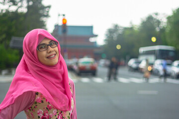Close up Portrait of beautiful young Asian muslim woman wearing hijab and eyeglasses in street of Kyoto. Face smiling at camera with happy expression. City traffic in bokeh background.