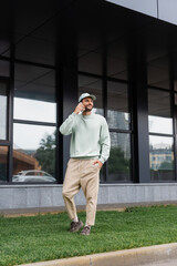 full length of pleased man standing with hand in pocket and talking on cellphone near modern building