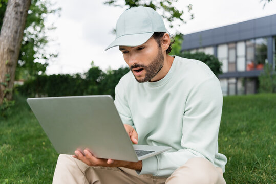 shocked freelancer in cap looking at laptop in park