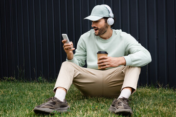 amazed man in cap listening music in wireless headphones and using cellphone while sitting on green...